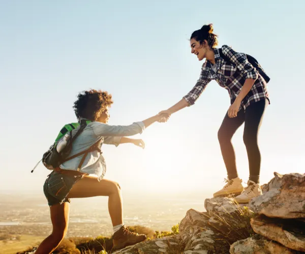 The image depicts two hikers on a mountain, one reaching out to help the other up a rocky incline. They are both wearing casual hiking attire and backpacks, and the setting sun casts a warm glow, highlighting their smiling faces and the expansive view in the background, suggesting a successful adventure or climb.